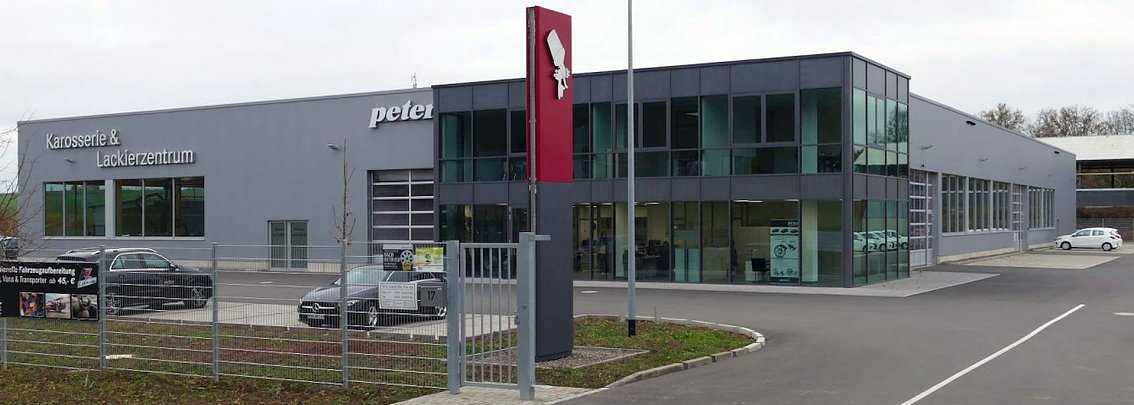 Modernes, grau gehaltenes Geb&auml;ude des Karosserie- und Lackierzentrums Peter in Heilbad Heiligenstadt mit gro&szlig;er Glasfassade. Davor stehen mehrere Autos, ein Zaun mit offenem Tor und ein rotes Schild mit Logo. Asphaltierte Zufahrt und heller Himmel im Hintergrund.