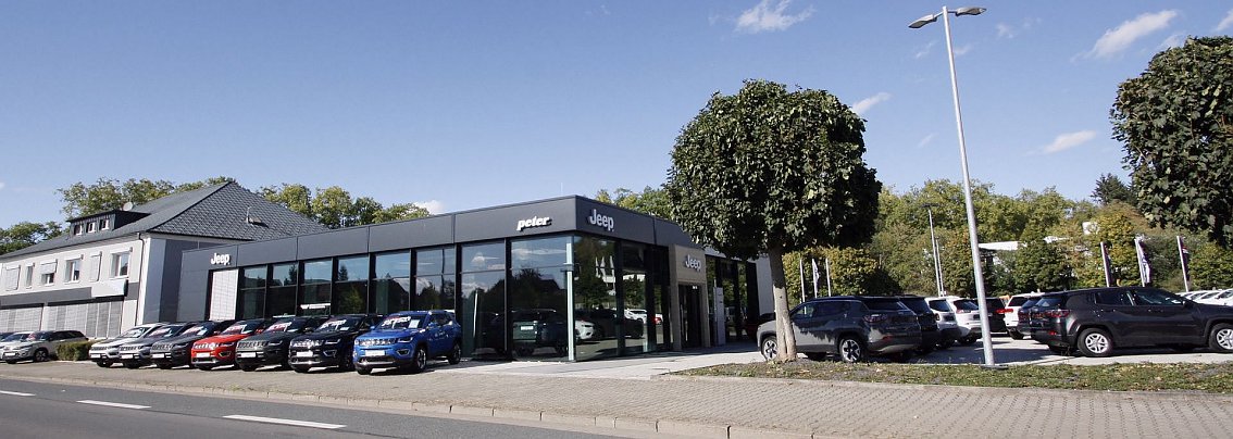 Peter-Autohaus mit gro&szlig;er Glasfassade in Nordhausen. Vor dem Geb&auml;ude stehen mehrere bunte Jeep-Modelle, daneben Parkpl&auml;tze mit weiteren Autos. Links ein graues Nebengeb&auml;ude, rechts B&auml;ume und eine Gr&uuml;nfl&auml;che. Blauer Himmel mit wenigen Wolken.