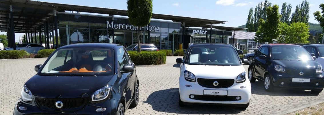 Reihe von Smart-Autos (schwarz und wei&szlig;) auf dem Parkplatz vor einem Mercedes-Benz Autohaus mit Peter-Logo in Northeim, im Hintergrund moderner Glasbau, gepflegte B&uuml;sche und B&auml;ume unter blauem Himmel.
