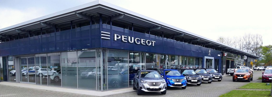Peugeot Autohaus in G&ouml;ttingen mit moderner Glasfassade und gro&szlig;em Peugeot-Logo, mehrere neue Peugeot-Fahrzeuge in verschiedenen Farben vor dem Geb&auml;ude geparkt.