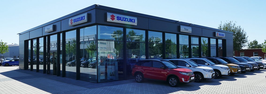 Modernes Suzuki Autohaus Peter in Erfurt mit gro&szlig;er Glasfassade, mehreren Suzuki Neuwagen in verschiedenen Farben (rot, wei&szlig;, gelb) vor dem Geb&auml;ude geparkt, sonniger Tag mit blauem Himmel.