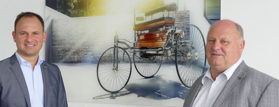 Andreas und Helmut Peter, Gesch&auml;ftsf&uuml;hrer der Autohaus-Peter-Gruppe, in Anz&uuml;gen stehen nebeneinander und l&auml;cheln in die Kamera; im Hintergrund ein gro&szlig;es Bild mit dem historischen Benz-Patent-Motorwagen.