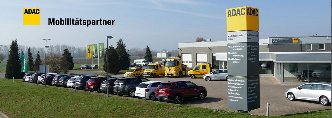 Au&szlig;enansicht des ADAC-Standorts Nordhausen an einem sonnigen Tag. Im Vordergrund eine Reihe geparkter Autos, dahinter ein gro&szlig;es graues Geb&auml;ude mit ADAC-Logo und gelber Beschilderung. Davor stehen mehrere gelbe ADAC-Pannenhilfefahrzeuge. Im Hintergrund Fahnen mit ADAC-Logo und gr&uuml;ner Wiese mit B&auml;umen.