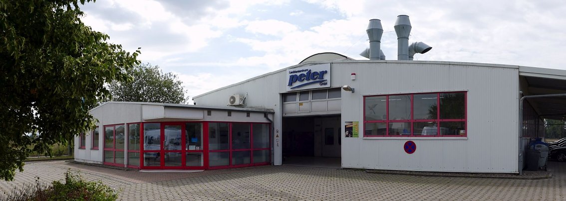 Eingangsbereich des Lackierzentrums Peter in Nordhausen. Flaches Geb&auml;ude mit wei&szlig;en Fassaden, roten Fensterrahmen und dem blauen Peter-Logo &uuml;ber dem Tor. Vor dem Geb&auml;ude gepflasterter Hof, links B&auml;ume und B&uuml;sche, auf dem Dach gro&szlig;e Abluftanlagen.