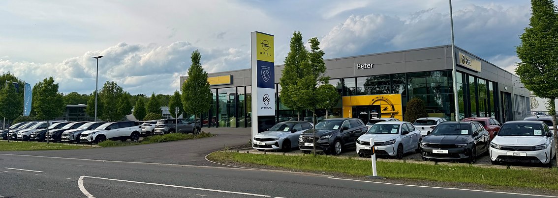 Modernes Nordh&auml;user Opel-Autohaus mit gro&szlig;er Glasfassade und grauer Verkleidung. Am Geb&auml;ude ist das Opel-Logo sowie der Schriftzug Peter sichtbar. Vor dem Eingang steht ein wei&szlig;er Opel-Transporter. Der Himmel ist klar und blau, es ist sonnig.