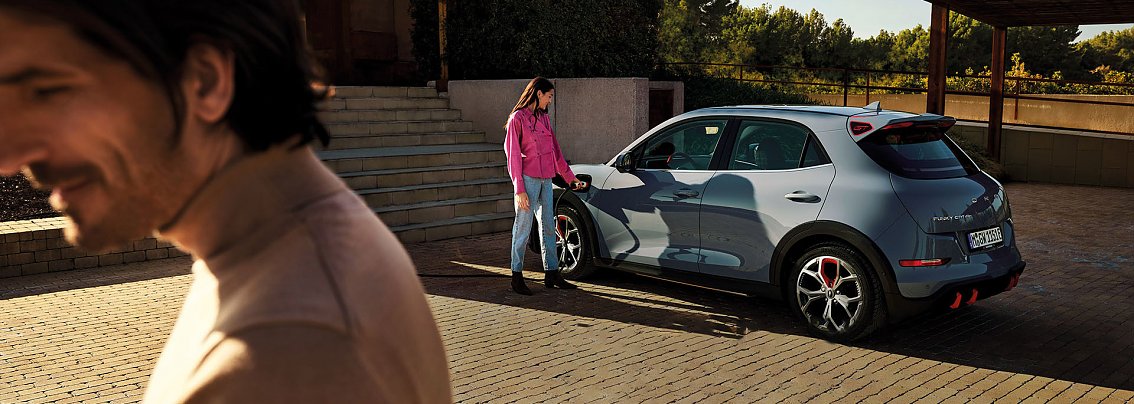 Eine Frau steht an einem grauen GWM Elektroauto und l&auml;dt es, im Vordergrund ein l&auml;chelnder Mann, sonnige und moderne Umgebung.