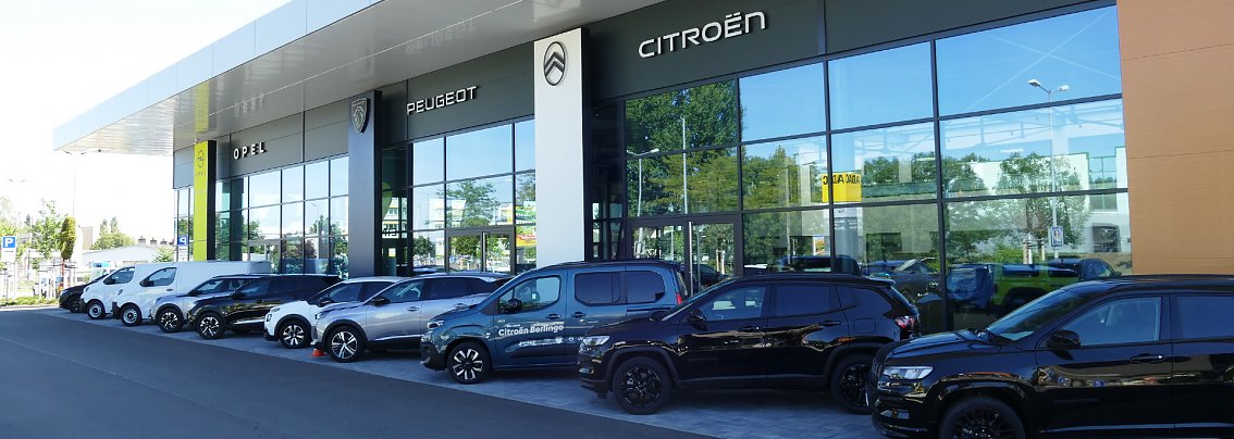 Moderne Autohaus-Fassade mit gro&szlig;en Glasfl&auml;chen und den Markenlogos von Opel, Peugeot und Citro&euml;n; davor eine Reihe geparkter Fahrzeuge in unterschiedlichen Farben (wei&szlig;, blau, schwarz), unter anderem Vans und SUVs.