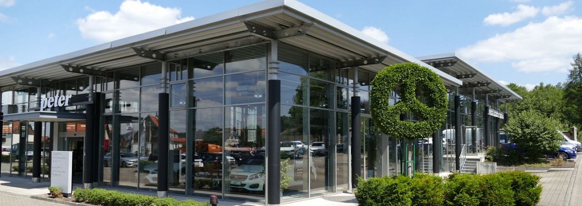 Modernes Mercedes-Benz Autohaus in Osterode mit gro&szlig;er Glasfassade. Vor dem Geb&auml;ude ist ein Mercedes-Stern aus gr&uuml;nem Buschwerk geformt. Mehrere Autos sind hinter der Glasfront sichtbar, davor gepflegte B&uuml;sche und blauer Himmel mit Wolken.