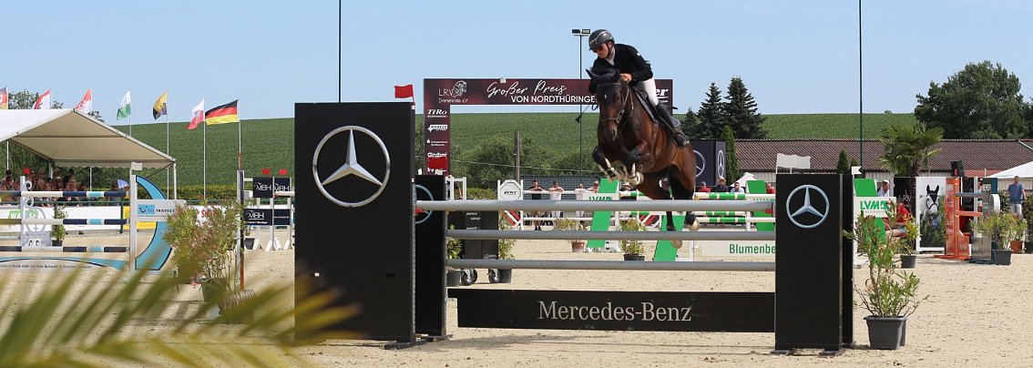 Das Bild zeigt einen Springreiter in Immenrode, der mit seinem Pferd &uuml;ber ein Hindernis mit Mercedes-Benz-Werbung springt. Im Hintergrund sieht man eine Reitturnier-Arena mit verschiedenen Hindernissen, Fahnen mehrerer L&auml;nder und Zuschauerbereichen. Das Wetter ist sonnig, und es gibt einige Pflanzen und Dekorationen um die Arena.