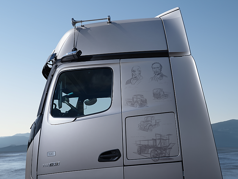 Silbernes Lkw-Fahrerhaus von der Seite vor blauem Himmel. Auf dem Lack hinter der Fensterscheibe sind feine Zeichnungen von historischen Autos und Portr&auml;ts von Carl Benz und Gottlieb Daimler.