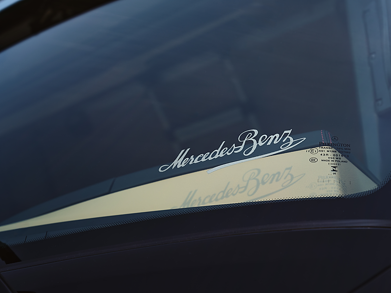 Nahaufnahme einer Windschutzscheibe. Im Glas ist der geschwungene, wei&szlig;e Schriftzug Mercedes-Benz in Schreibschrift eingelassen. Darunter wirft er einen Schatten auf das Armaturenbrett. Rechts stehen kleine Pr&uuml;fzeichen auf dem Glas.