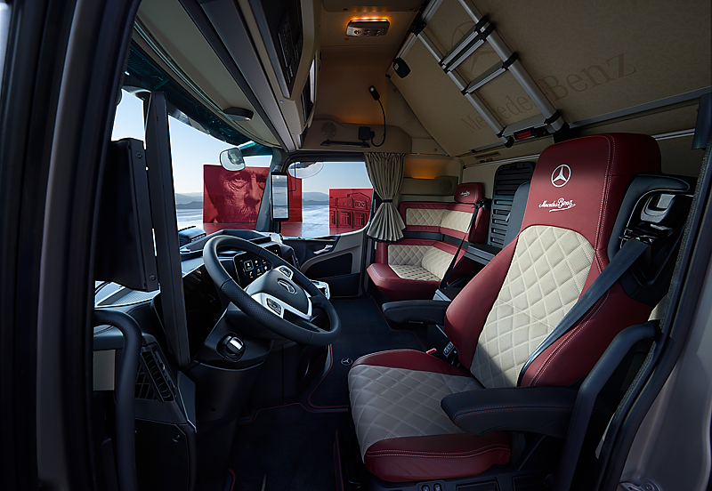 Moderner Lkw-Innenraum von vorne links fotografiert. Schwarzes Lenkrad mit Kn&ouml;pfen im Vordergrund. Beigefarbene Ledersitze mit roter Umrandung und Mercedes-Logo. Im Hintergrund eine gem&uuml;tliche Sitzecke. 