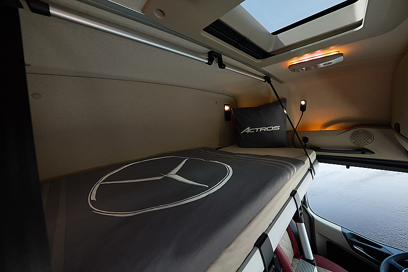 Schlafkabine im Lkw-Fahrerhaus. Ein schmales Bett mit grauer Decke, auf der ein gro&szlig;er Mercedes-Stern prallt. Am Kopfende liegt ein graues Kissen mit dem Schriftzug Actros. Oben eine kleine Dachluke und warmes Licht aus einer Lampe.