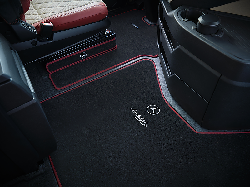 Schwarzer Lkw-Fu&szlig;boden im Detail. Die passgenauen Teppiche haben eine auff&auml;llige, rote Umrandung. Auf dem Teppich im Vordergrund sind das wei&szlig;e Mercedes-Benz-Logo und der geschwungene Markenschriftzug aufgestickt.