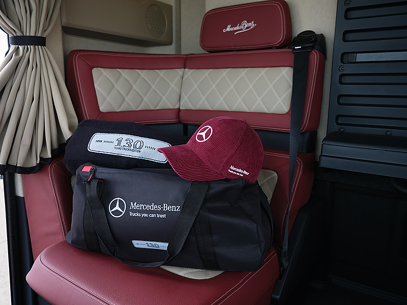 Fan-Artikel auf einem roten Lkw-Sitz. Eine gro&szlig;e, schwarze Reisetasche mit wei&szlig;em Mercedes-Benz Logo liegt auf der Sitzfl&auml;che. Darauf liegen eine dunkelrote Baseballkappe mit wei&szlig;em Stern und eine gefaltete, schwarze Decke mit der Zahl 130.