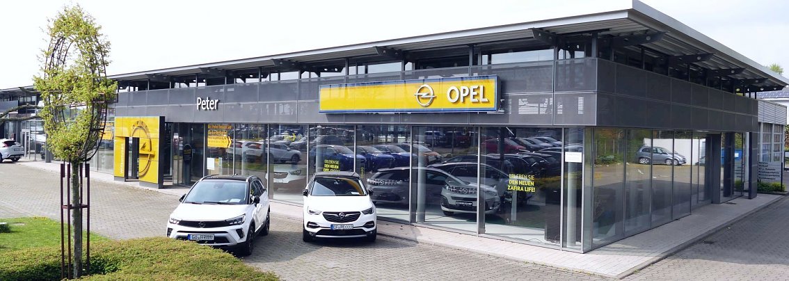 Au&szlig;enansicht eines modernen Opel-Autohauses in G&ouml;ttingen. Das Geb&auml;ude hat eine gro&szlig;e Glasfassade mit gelbem Opel-Schriftzug und Logo. Vor dem Eingang stehen zwei wei&szlig;e Opel-Fahrzeuge auf dem Parkplatz. Links ist ein gestalteter Baum in Form des Opel-Logos zu sehen. Das Autohaus wirkt hell und einladend, der Himmel ist leicht bew&ouml;lkt.