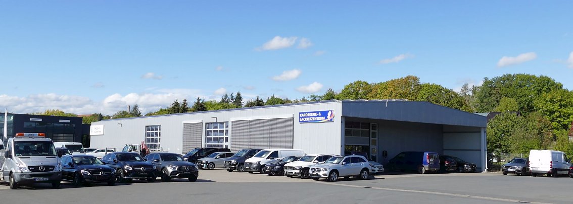 Graues Geb&auml;ude mit dem Schild �Karosserie & Lackierzentrum� in Nordhausen. Davor sind zahlreiche Autos, darunter mehrere Mercedes-Modelle und Transporter, auf dem Parkplatz abgestellt. Im Hintergrund gr&uuml;ne B&auml;ume und blauer Himmel mit wenigen Wolken.