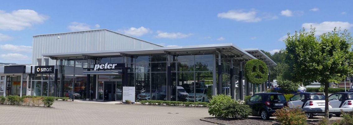 Moderne Glasfassade des Autohauses Peter in Osterode. &Uuml;ber dem Eingang sind die Logos von Smart und Peter angebracht. Vor dem Geb&auml;ude stehen mehrere Autos, daneben Gr&uuml;nfl&auml;chen mit B&uuml;schen und B&auml;umen. Der Himmel ist blau mit vereinzelten wei&szlig;en Wolken.