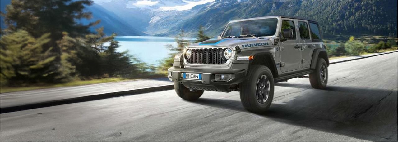 Grauer Jeep Wrangler Rubicon f&auml;hrt auf einer Stra&szlig;e an einem See vorbei, umgeben von Bergen und W&auml;ldern im Hintergrund.