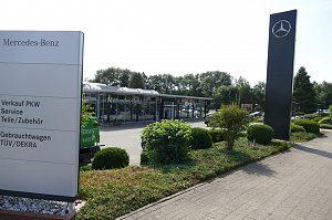 Außenansicht des Mercedes-Benz Autohauses Duderstadt mit großem Mercedes-Stern auf einer hohen Stele und einem Wegweiser, der auf Verkauf PKW, Service, Teile/Zubehör, Gebrauchtwagen sowie TÜV/DEKRA hinweist. Im Hintergrund ein modernes, verglastes Autohausgebäude, umgeben von gepflegten Grünflächen und Büschen.