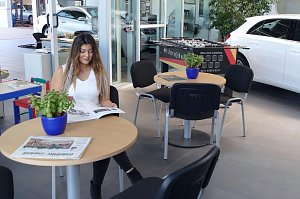 Wartebereich des Duderstädter Mercedes-Autohauses mit einer jungen Frau, die eine Zeitschrift liest, mehreren Tischen mit Zeitungen und Pflanzen, einem Kicker-Tisch im Hintergrund sowie einem weißen Auto, das durch eine Glaswand sichtbar ist.