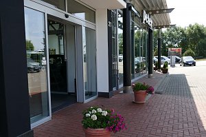 Eingangsbereich eines Autohauses der Peter Gruppe in Duderstadt, mit großen Glasfassaden, schwarzer Beschilderung und Blumen in Töpfen entlang des gepflasterten Weges; rechts stehen geparkte Autos, im Hintergrund Bäume und ein Parkplatz.