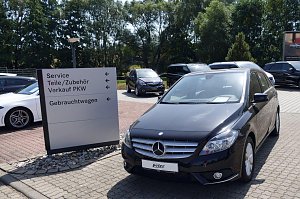 Parkplatz der Mercedes-Benz-Niederlassung in Duderstadt mit einem schwarzen Mercedes-Benz im Vordergrund. Daneben ein weißes Auto und ein Wegweiser mit Beschriftungen wie Service, Teile/Zubehör, Verkauf PKW und Gebrauchtwagen. Im Hintergrund mehrere weitere Fahrzeuge und Bäume.