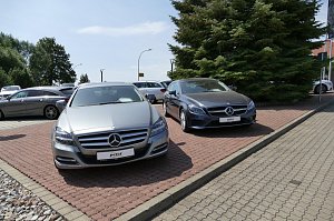 Zwei Mercedes-Benz Fahrzeuge stehen nebeneinander auf einem gepflasterten Parkplatz. Links ein silberner Mercedes und rechts ein dunkler Mercedes. Im Hintergrund weitere Fahrzeuge und große grüne Tannenbäume.