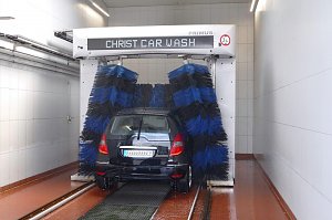 Auto in einer automatischen Waschanlage. Blaue und schwarze Bürsten reinigen das schwarze Fahrzeug. Die Umgebung ist hell und gekachelt, mit rutschfestem Boden.
