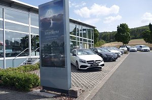 Reihe von Mercedes-Benz Gebrauchtwagen (Junge Sterne) auf dem Parkplatz eines Autohauses. Im Vordergrund steht ein großes Schild mit dem Logo und der Aufschrift �Junge Sterne � Die besten Gebrauchten von Mercedes-Benz�. Im Hintergrund moderne Glasfassade des Autohauses, Bäume und Felder bei sonnigem Wetter.