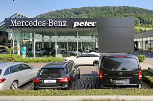 Mercedes-Benz Autohaus Peter mit Glasfassade und großem Logo, davor geparkte Mercedes-Fahrzeuge, im Hintergrund grüne bewaldete Hügel und blauer Himmel.