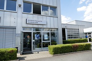 Eingangsbereich eines Autohauses mit Glasfront. Über der Tür hängt ein Schild mit der Aufschrift �Annahme Transporter, Annahme LKW�. Rechts ist eine Werkstatthalle mit großen Toren sichtbar, davor gepflegte Hecken und ein gepflasterter Weg.