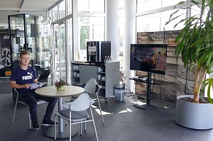 Heller Wartebereich im Mercedes-Autohaus mit Glasfront. Ein junger Mann sitzt an einem runden Tisch und liest eine Broschüre. Neben ihm stehen weitere Stühle, ein kleiner Tisch mit einer Pflanze, ein Kaffeeautomat mit Tassenregal und ein großer Fernseher. Rechts im Bild eine große grüne Pflanze in einem grauen Topf.