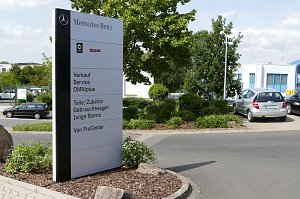 Großes weißes Schild auf dem Gelände eines Mercedes-Benz Autohauses. Auf dem Schild stehen Logos von Mercedes-Benz und Smart sowie Hinweise auf angebotene Leistungen: Verkauf, Service, OMNIplus, Teile/Zubehör, Gebrauchtwagen, Junge Sterne und Van ProCenter. Im Hintergrund sieht man geparkte Autos, grüne Sträucher und Gebäude.