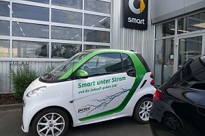 Weiß-grüner Smart mit der Aufschrift �Smart unter Strom und die Zukunft gehört DIR!�, geparkt vor einem Autohaus. Auf dem Auto ist ein Blitzmotiv abgebildet. Im Hintergrund ist das Smart-Logo an der Gebäudewand zu sehen.