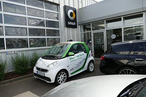 Weiß-grüner Smart mit der Aufschrift �Smart unter Strom und die Zukunft gehört DIR!�, geparkt vor einem Autohaus. Auf dem Auto ist ein Blitzmotiv abgebildet. Im Hintergrund ist das Smart-Logo an der Gebäudewand zu sehen.