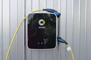 Ladestation mit Smart-Logo an einer silberfarbenen Metallwand. Die Station hat ein schwarzes Frontpanel mit gelbem Kabel und blau-schwarzem Stecker, der an der Seite hängt.