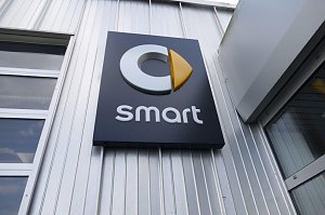 Nahaufnahme eines schwarzen Schildes mit dem Smart-Logo an einer silberfarbenen Metallfassade. Das Logo besteht aus einem stilisierten grauen �C� mit einem gelben, pfeilförmigen Element, darunter steht der Schriftzug �smart� in weißen Buchstaben.