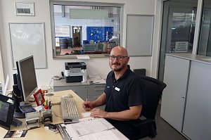 Freundlich lächelnder Mitarbeiter mit Brille und kurzärmeligem Poloshirt sitzt an einem Schreibtisch in einem Autohaus-Büro. Vor ihm liegen Dokumente, ein Computer, Tastatur und Büromaterial. Im Hintergrund ist durch ein Fenster eine Werkstatt mit Reifenstapeln und Werkzeugen sichtbar.
