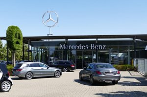Außenansicht eines Mercedes-Benz-Autohauses mit großem Schriftzug �Mercedes-Benz� an der Glasfassade. Darüber ein großes silbernes Mercedes-Logo auf dem Dach. Vor dem Gebäude stehen mehrere silberne und schwarze Mercedes-Fahrzeuge. Links eine grüne, in Form des Markenlogos geschnittene Buchsbaumfigur.