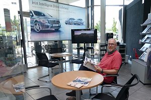 Heller Wartebereich in einem Mercedes-Benz-Autohaus. Ein Mann sitzt an einem runden Tisch, liest eine Broschüre und schaut in die Kamera. Neben ihm liegen Magazine auf dem Tisch. Im Hintergrund ein großer Werbebanner mit Mercedes-Autos, ein Fernseher, Stühle, ein Kaffeeautomat und Regale mit Prospekten.