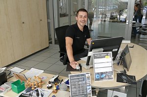 Junger, freundlich lächelnder Mitarbeiter sitzt an einem Schreibtisch im Autohaus. Er trägt ein schwarzes Poloshirt mit Mercedes-Logo. Vor ihm stehen ein Computer, viele Unterlagen, Stempel, Prospekte und Schilder mit Service-Angeboten. Im Hintergrund Glastüren und ein heller Empfangsbereich.