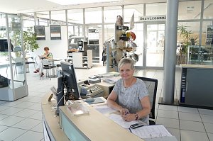 Freundliche Mitarbeiterin sitzt an einem Empfangstresen im Autohaus, vor ihr ein Computer, Telefon und Unterlagen. Sie lächelt in die Kamera. Im Hintergrund sitzt eine weitere Frau an einem Tisch und liest. Helle, moderne Empfangshalle mit Glaswänden, Pflanzen und Schildern, auf einem Glastür-Bereich steht �Serviceannahme�.