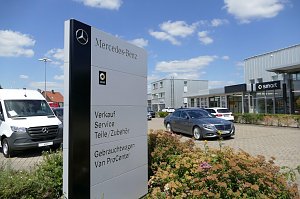 Schild mit Mercedes-Benz- und Smart-Logo vor einem modernen Autohaus mit Glasfassade. Auf dem Schild stehen Angebote wie Verkauf, Service und Gebrauchtwagen. Davor parken ein grauer Mercedes-Pkw und ein weißer Mercedes-Van. Blumenbeet im Vordergrund, blauer Himmel mit wenigen Wolken im Hintergrund.