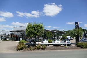 Autohaus mit großer Glasfassade und den Logos von Smart und Peter. Auf dem Vorplatz sind mehrere geparkte Smart-Autos zu sehen. Rechts steht ein hoher schwarzer Mast mit dem Mercedes-Benz-Stern. Vor dem Gebäude befinden sich Bäume und Büsche. Der Himmel ist blau mit vereinzelten Wolken.