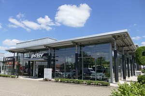 Modernes Mercedes-Benz Autohaus mit großer Glasfassade und der Aufschrift �peter� über dem Eingang. Links daneben ein Bereich mit Smart-Logo. Vor dem Gebäude gepflegte Grünflächen, blauer Himmel mit wenigen weißen Wolken.