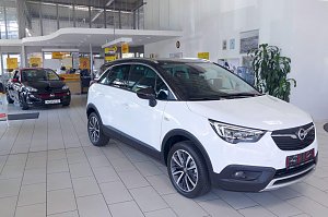 Im Innenraum eines Autohauses stehen zwei Opel-Fahrzeuge. Im Vordergrund ein weißer Opel Crossland mit schwarzen Akzenten, im Hintergrund ein schwarzer Opel Adam mit rotem Dach. Der helle Ausstellungsraum ist modern gestaltet, mit großen Fenstern und einem Verkaufsbereich im Hintergrund. Der Boden ist hell gefliest.