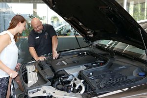 Ein Mercedes-Benz Servicemitarbeiter erklärt einer jungen Frau den Motorraum eines Fahrzeugs. Die Motorhaube ist geöffnet, beide schauen gemeinsam in den Motor. Im Hintergrund helle Glasfronten des Autohauses.