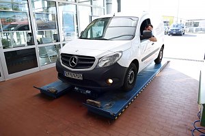 Ein weißer Mercedes-Transporter mit Fahrer am Steuer steht in der Autohaus-Werkstatt auf einem blauen Prüfstand, bereit zur Inspektion.