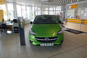 Heller Verkaufsraum des Opel-Autohauses mit einem gr&uuml;nen Opel Corsa im Vordergrund. Links stehen Schreibtische mit St&uuml;hlen und Informationsmaterialien. Im Hintergrund sieht man den Empfangsbereich, an der eine Mitarbeiterin sitzt. Gro&szlig;e Fenster lassen viel Tageslicht herein, und die Decke ist mit wei&szlig;en Paneelen versehen. Ein Schild mit der Aufschrift "CORSA" steht neben dem Auto.